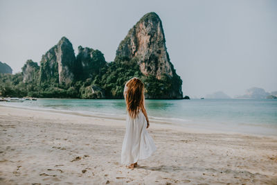 Back view full body of faceless female in white dress standing on sandy beach near azure water against rocky cliffs in thailand