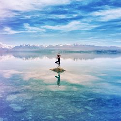 Full length of woman standing on one leg over rock in lake against sky