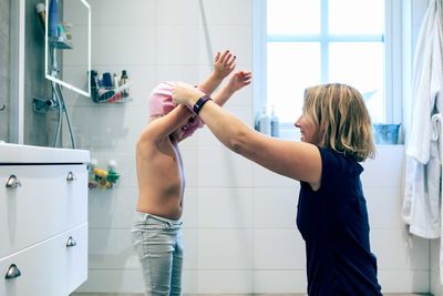 Side view of smiling mother removing daughter's clothes in bathroom