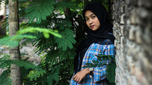 Portrait of young woman standing by plants against stone wall