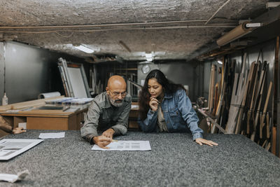 Mature bald owner discussing blueprint with female client at workbench