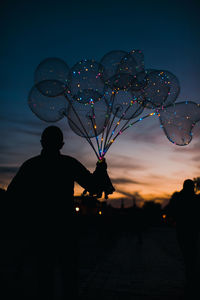 Rear view of silhouette man holding illuminated balloons against sky during sunset