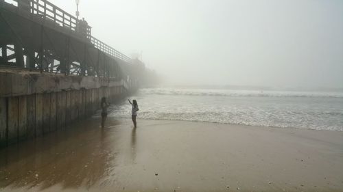 Women by pier at beach against sky during foggy weather