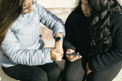 Hispanic mother  checking time on smartwatch with daughter using smartphone in park on sunny day