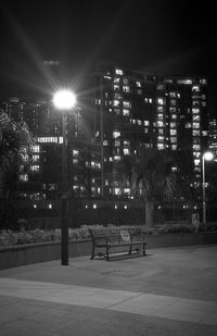 Empty bench by illuminated buildings against sky at night