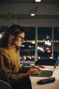 Side view of female professional working late while using laptop at illuminated desk in office