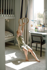 Full length of happy boy hanging from gymnastic rings in bedroom at home