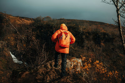 Back view of young tourist in winter wear looking at cascade and mountain river between stone hill