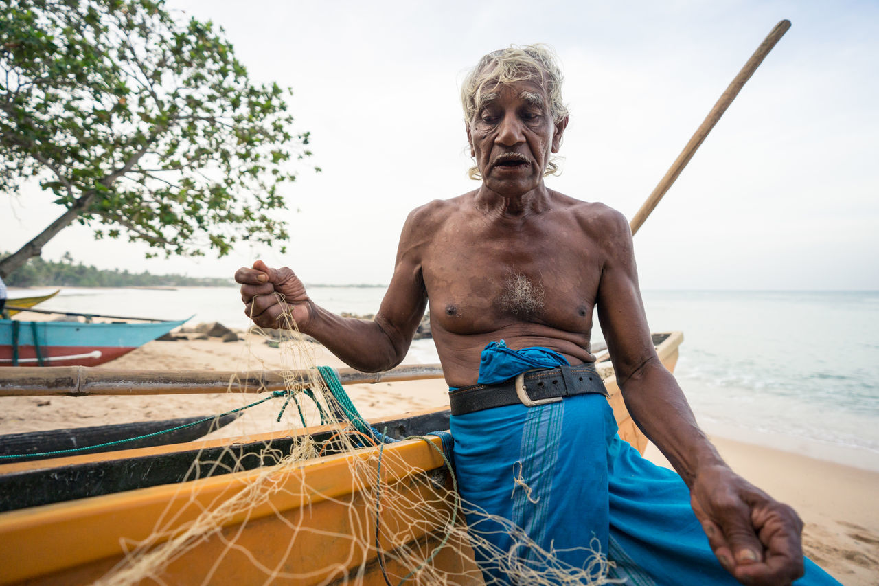 Tangalle, sri lanka - july 28, 2019: ethnic old fishermen in casual light wear unwinding fishing net on big yellow boat at tropical sandy beach