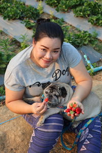 High angle portrait of woman feeding strawberry to dog while sitting in farm