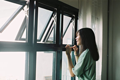 Side view of woman using phone while standing on window