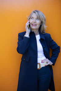 Confident woman talking on the phone against a vibrant orange wall in a stylish outfit