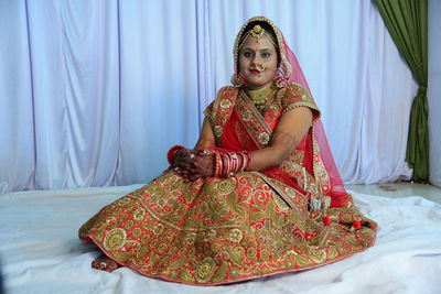 Full length of bride sitting on sheet against curtains