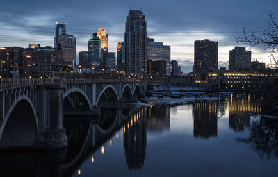 Bridge over river by illuminated buildings against sky at dusk