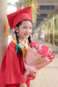 Smiling girl in graduation gown holding flowers while standing outdoors