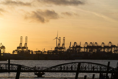 Silhouette cranes at harbor against sky during sunset