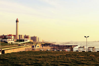 Sea by buildings and lighthouse at sunset