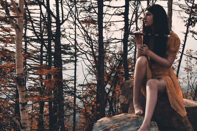 Young woman holding flute while sitting on rock in forest during autumn