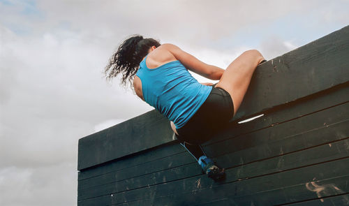 Low angle view of sporty woman climbing wooden wall against sky