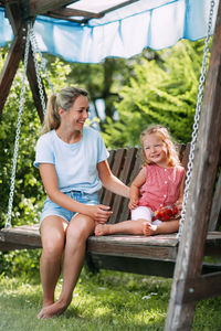 A happy mother is sitting on a swing with her charming daughter, who is eating fresh strawberries. 