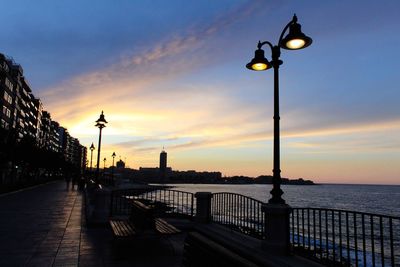 Illuminated street light by sea against sky during sunset