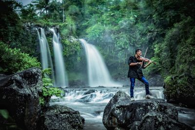 Man playing violin while standing on rock against waterfall in forest