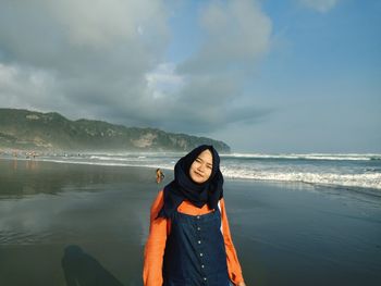 Portrait of young woman standing at beach against cloudy sky