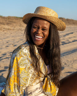 Cheerful african american female in beachwear and sunhat looking away and laughing at joke while spending sunny summer day on beach