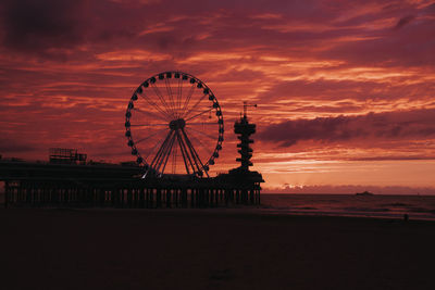 Silhouette ferris wheel by sea against orange sky