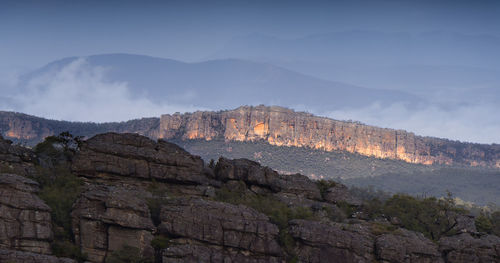 Scenic view of sunlit rocky mountains against sky with low wispy cloud