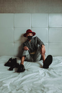 Full length of young man sitting on bed against wall