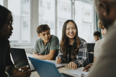Smiling teenage girl discussing with male teacher while sitting with friends in classroom