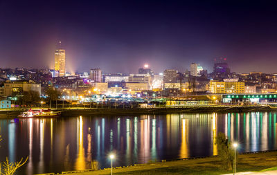 Illuminated buildings by river against sky at night