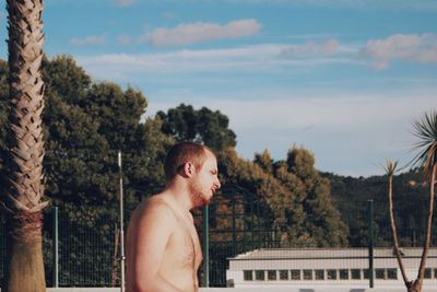 Side view of shirtless young man standing by palm trees against sky