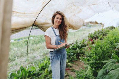 Happy farm worker with clipboard taking vegetables inventory at greenhouse