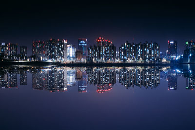 Illuminated modern buildings by sea against sky at night