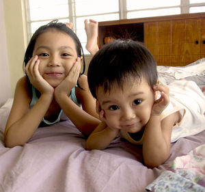 Close-up portrait of siblings with hands on cheeks lying on bed at home