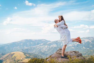 Male partner embracing female while standing on mountain against cloudy sky