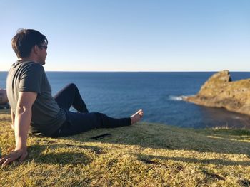 Man sitting on rock looking at sea against clear sky