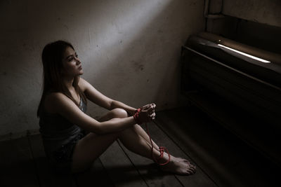 Side view of young woman with tied hands and legs sitting on floor at home