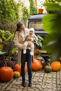 Woman playing with daughter while standing by pumpkins against trees
