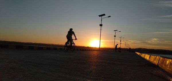 Silhouette man riding bicycle on street against sky during sunset