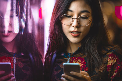 Smiling young woman using smart phone by window with reflection at night 