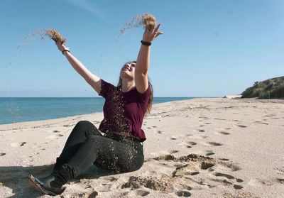 Young woman throwing sand while sitting on beach against sky