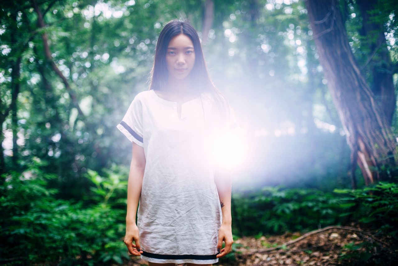 young adult, lifestyles, tree, leisure activity, forest, front view, person, casual clothing, young women, portrait, looking at camera, sunlight, sunbeam, standing, lens flare, focus on foreground, three quarter length