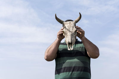 Young man holding a cow skull in front of his head in front of blue sky, low angle view point