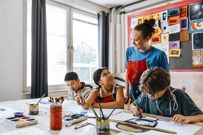 Smiling female teacher standing near students painting while sitting in classroom at school