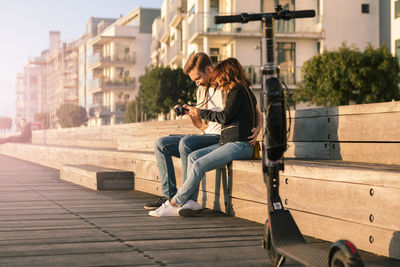 Loving couple sitting on seat while looking at camera during summer vacation in city