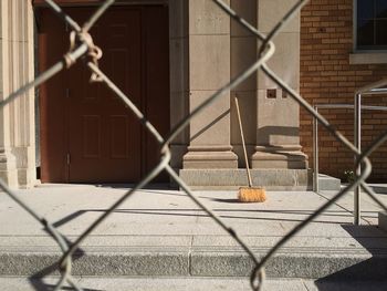 Broom on sidewalk against building seen through chainlink fence