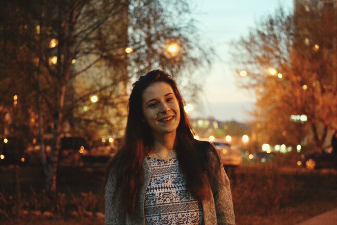 PORTRAIT OF SMILING YOUNG WOMAN STANDING IN PARK